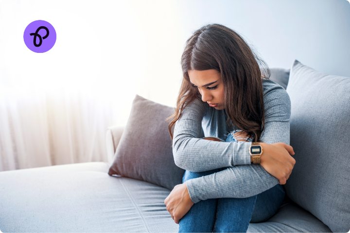 A teenage girl is sat on a bed, she wears a grey top and her bedding is grey. She has her knees up to her chest and her arms around her knees. She looks upset.