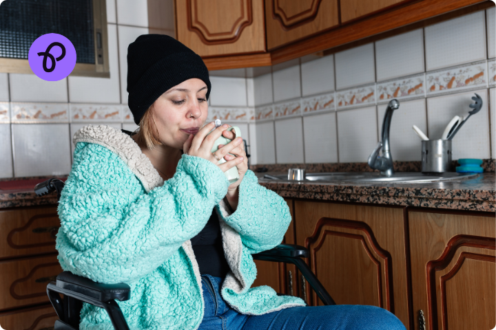 A woman is using a wheelchair, she is wearing a fleece top and a wooly hat, she is in a kitchen drinking a hot drink for a blog about benefits for disabled people to keep warm in winter