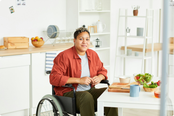 A person has short dark hair with lighter curls and wears a red shirt, grey top and trousers and sits in a wheelchair at a kitchen table 