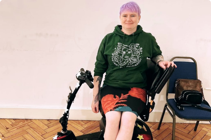 Keziah, a woman with short purple hair, sits in a mobility scooter and gazes frankly at the camera, an enigmatic smile on her face. She wears a floral patterned business skirt and a hoodie with a picture of Medusa and the legend “Petrify the Patriarchy”.