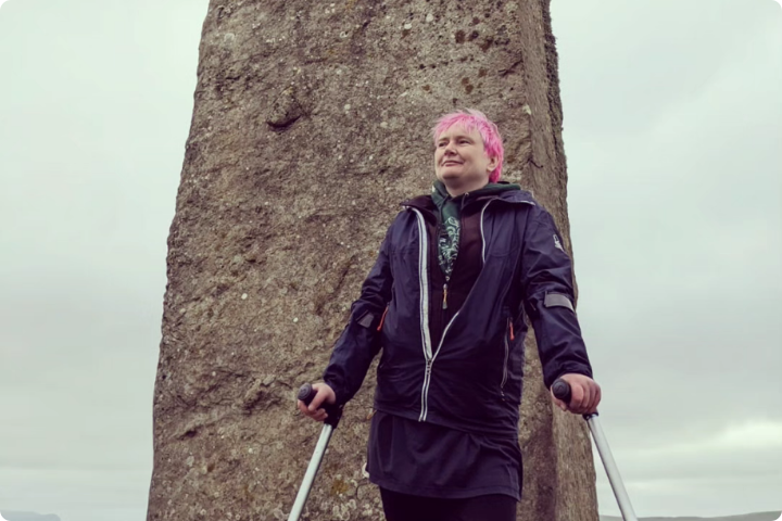 Keziah, a woman in her early forties with short pink hair, gazes into the distance in front of a large standing stone against a hazy grey sky. She is dressed for the outdoors and supported by a pair of crutches.