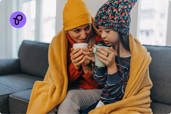 A woman and a little girl sit on a sofa wearing wooly hats and wrapped together in a yellow blanket whilst drinking hot drinks from mugs in a blog article about how to keep warm in winter for disabled people