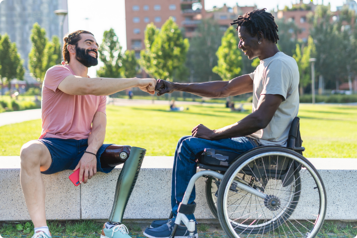 Two men sit on a wall in a park in a city, one man has dark hair and a beard and is wearing a pink t-shirt and blue shorts, he has a green metal prosthetic leg, the second man is black and has short plaited hair, a grey tshirt, blue jogging bottoms and trainers and is a wheelchair user