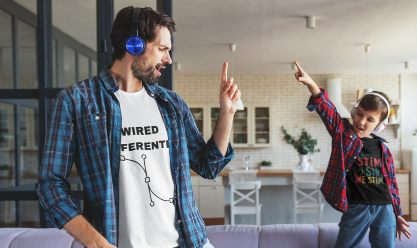 A dad and a pre teen son are wearing tshirts that promote acceptance of neurodiversity and checked shirts and headphones and are dancing in a kitchen