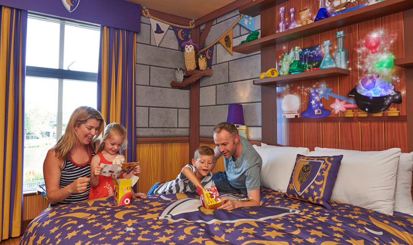 A Harry Potter style hotel room with potions and wizards hats on the wall and a family with a mum, dad and two young children on the bed having fun