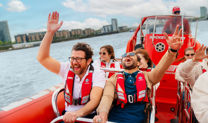 A red speed boat is rushing down a city river, there are a group of people on board but we focus on two white men in the foreground throwing their arms in the arm and laughing wildly