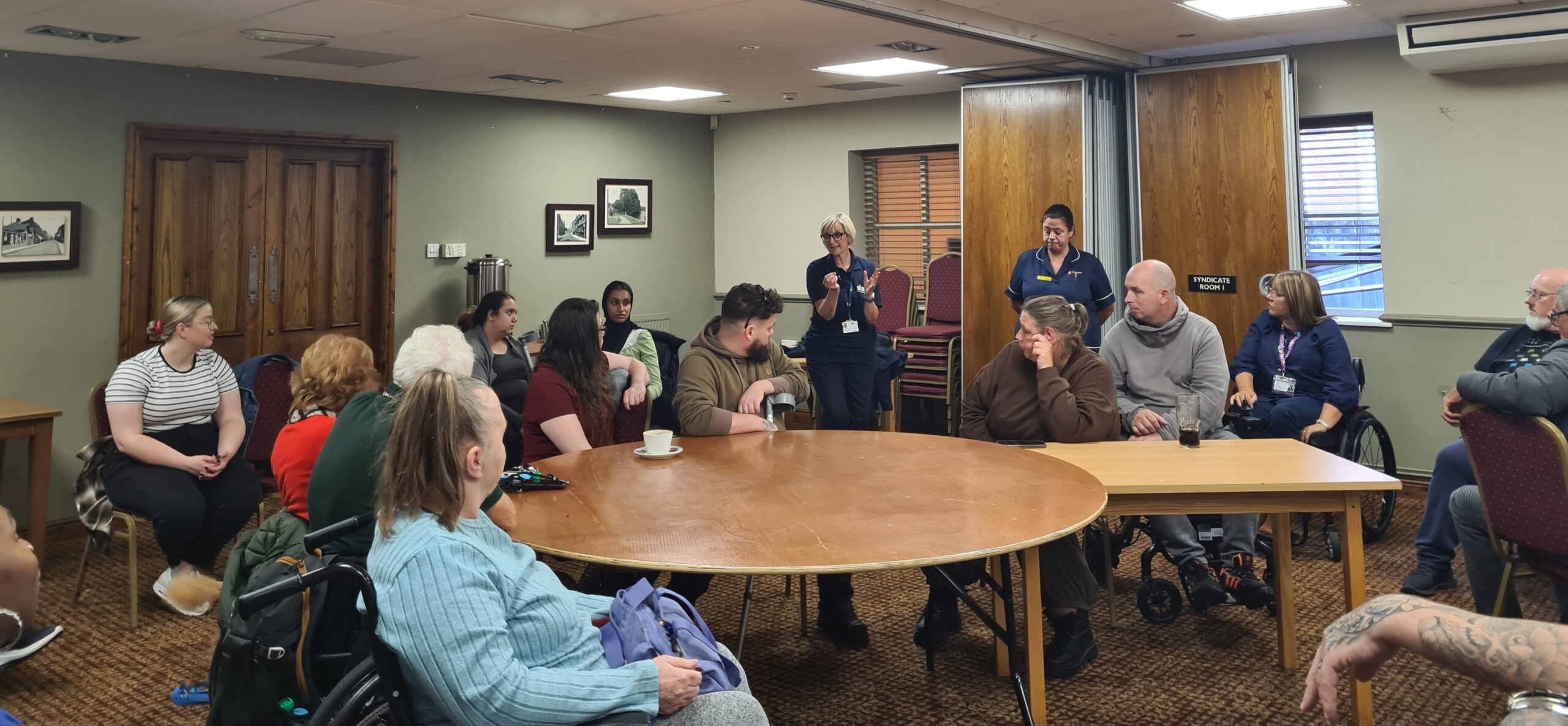 A group of smiling individuals, including wheelchair users, gathered in a meeting room from the spinal injuries Association for Purpl disabled discounts