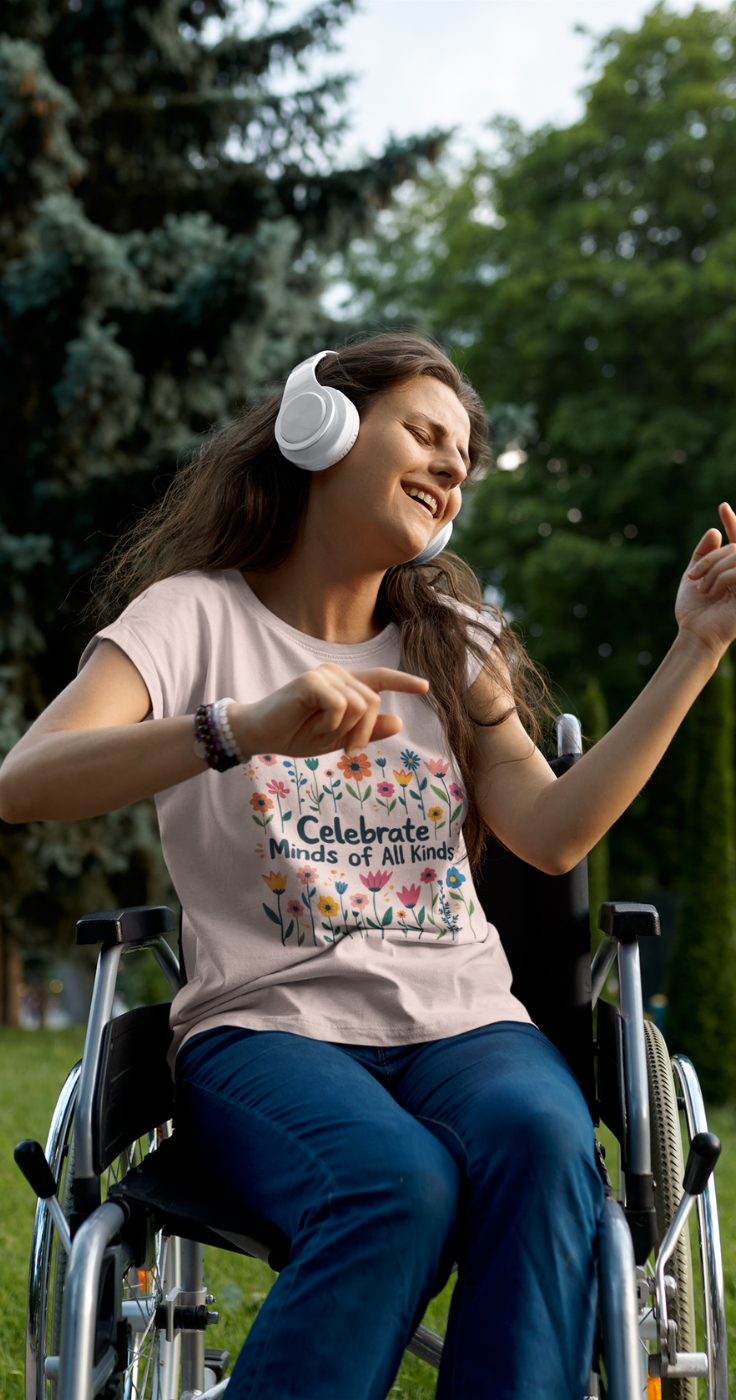 A young woman sits in a wheelchair with headphones on and is dancing, she is wearing a pink differently perfect t-shirt that says celebrate minds of all kinds and lots of flowers on it