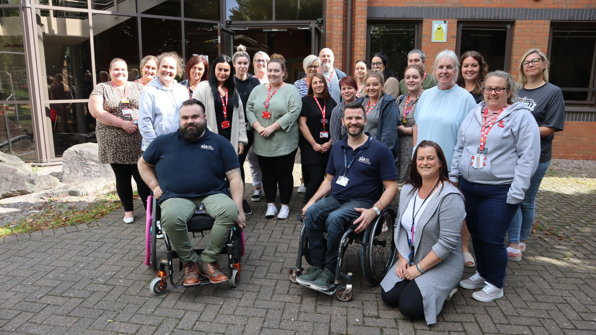 A group of smiling individuals, including wheelchair users, gathered in front of a building from the spinal injuries Association for Purpl disabled discounts