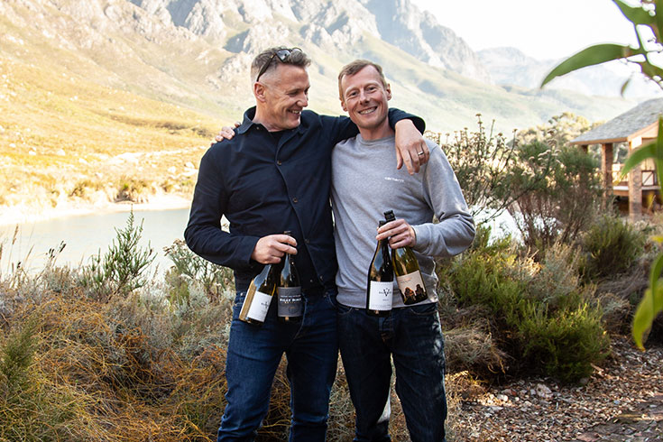 Two men holding bottles of Virgin Wines in a scenic vineyard, showcasing premium and sustainable selections.