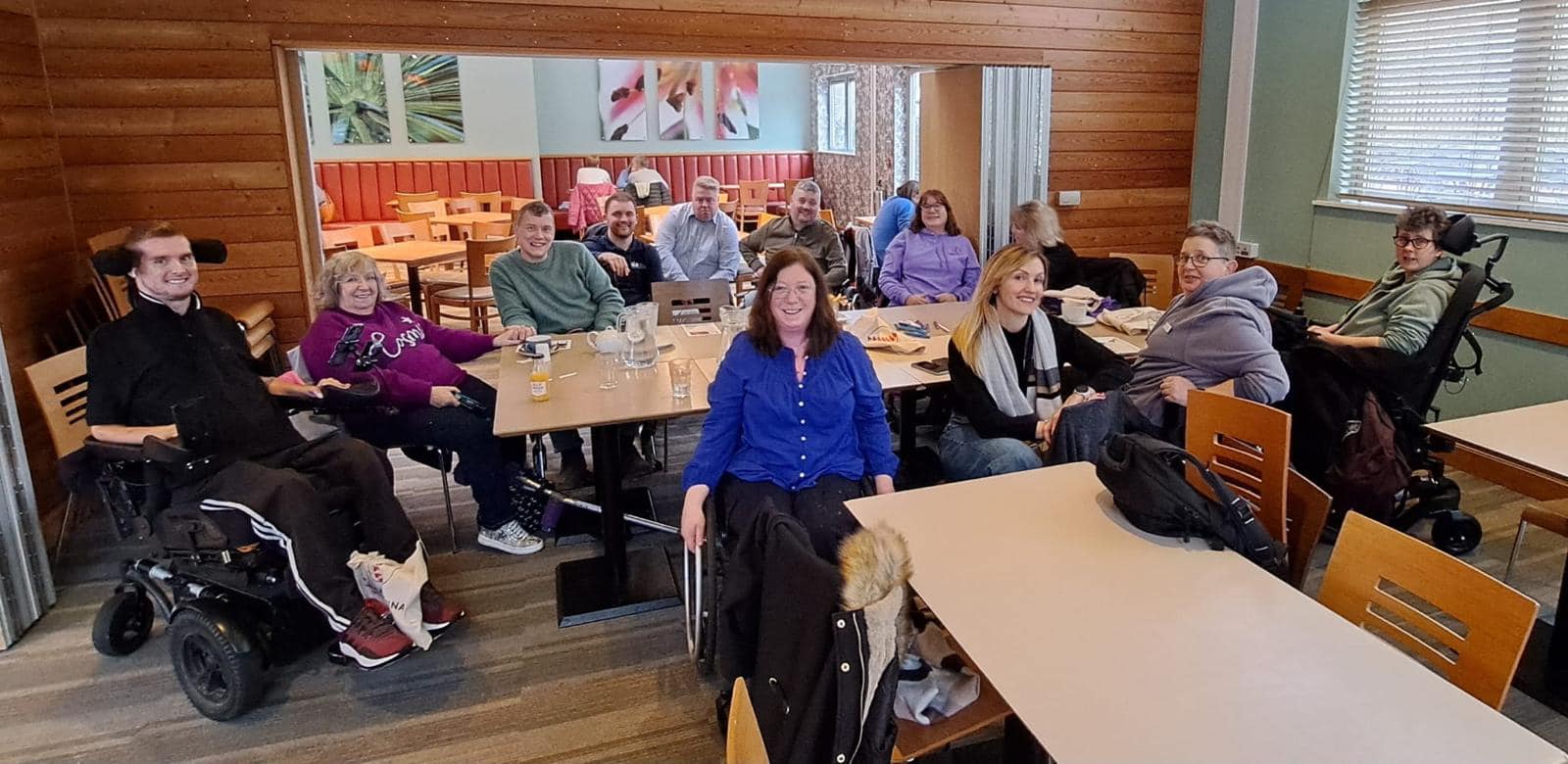 Community group of smiling individuals, including wheelchair users, gathered in a cafe from the spinal injuries Association for Purpl disabled discounts