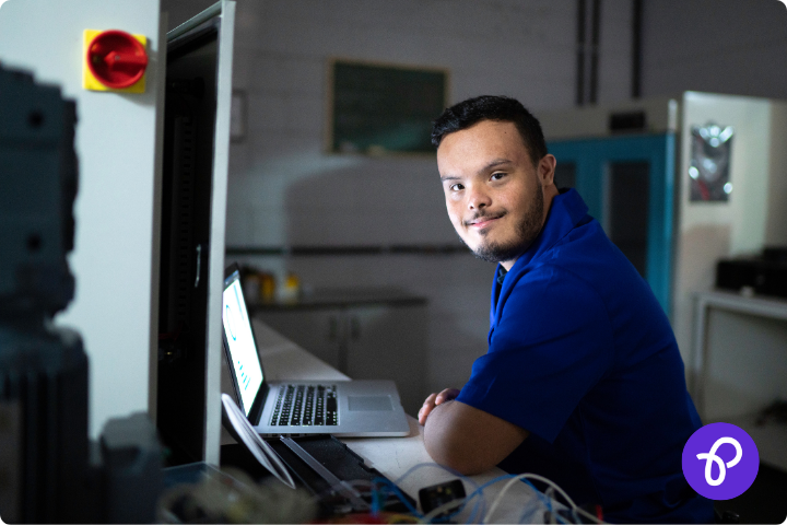 Young disabled man sits at a laptop at work, he is wearing a blue polo shirt and is smiling to camera, the background looks like a factory or workroom, for a blog about the get Britain working white paper and how it affects disabled people