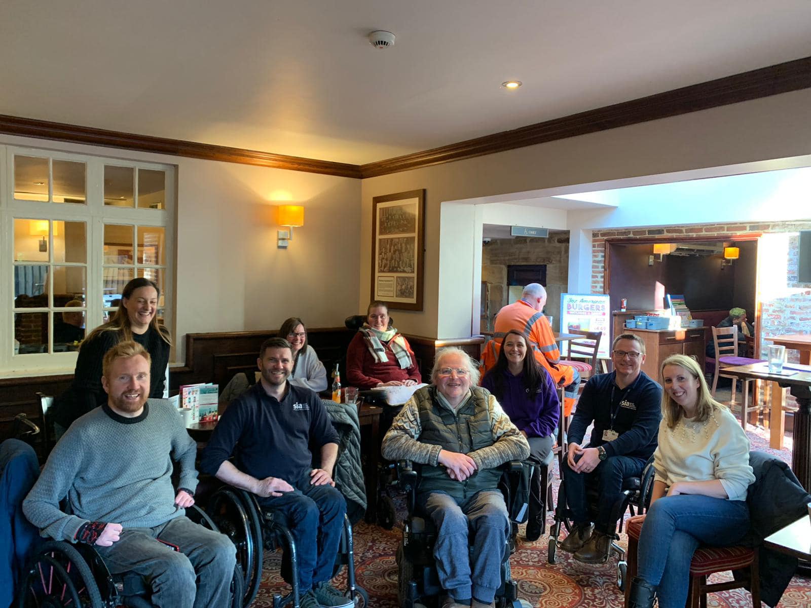 A group of smiling individuals, including wheelchair users, gathered in a cafe from the spinal injuries Association for Purpl disabled discounts