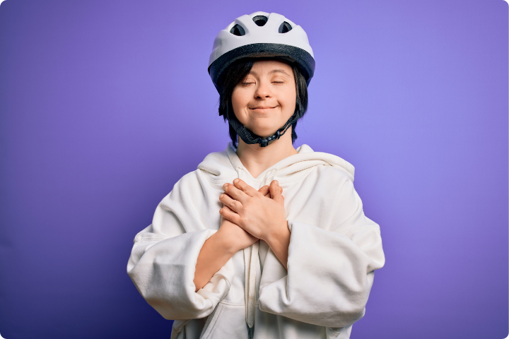 A white person with Down's Syndrome is wearing a white hoody and white cycling helmet, they have their eyes closed with a calm relaxed smile on their face and their hands clasped over their chest. They look serene and unstressed. There is a purple plain background 