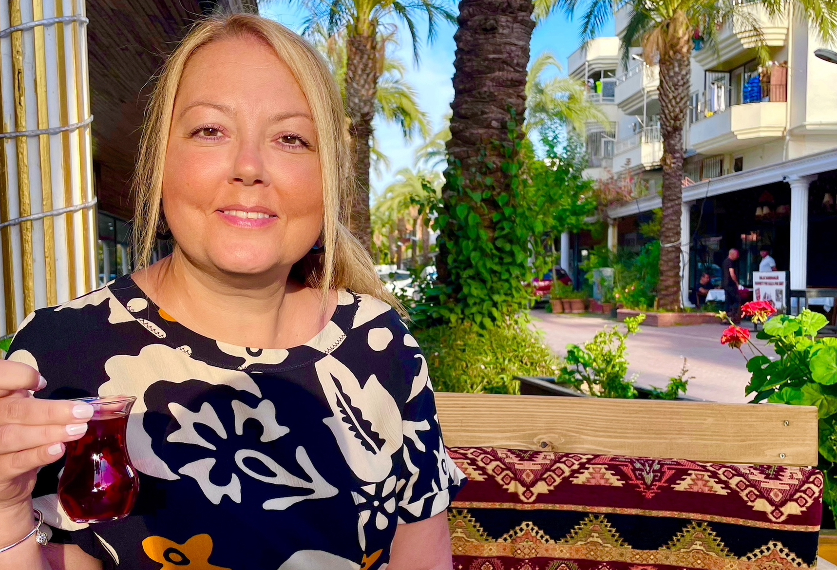 Christy is a blonde white woman aged 39, she is smiling to camera wearing a black and white patterned top and is sat on a bench, it looks like she is on holiday as there are palm trees and a concourse behind her. She is holding a glass with red/purple liquid in it as though she is about to cheers someone and is smiling and looks relaxed and content.