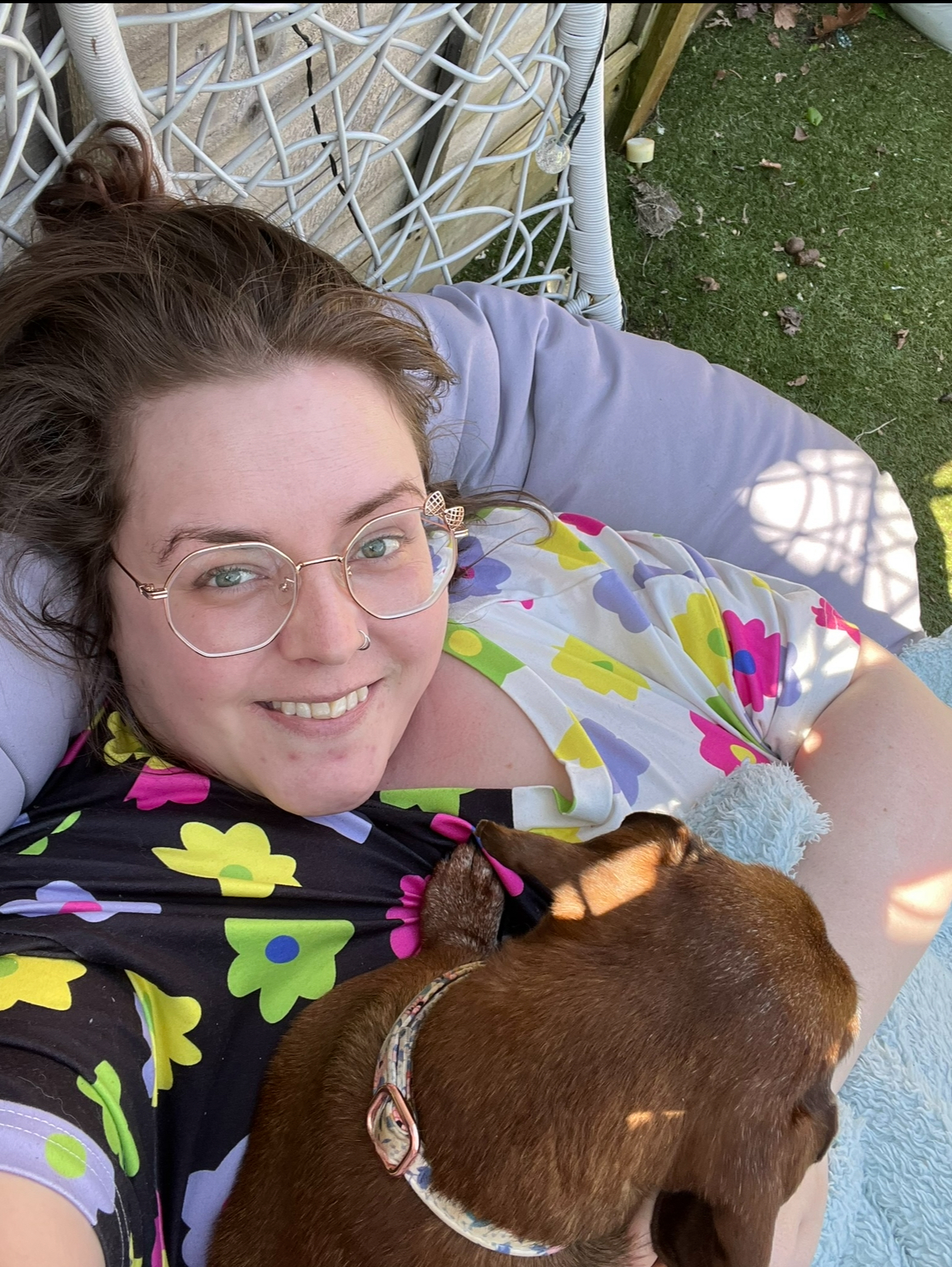 Rebecca is laid on a purple cushion outdoors on grass, she has a dog sat on her and she is wearing a colourful flowery top, gold rimmed glasses with a bow on them, she has brown hair and a nose ring and is smiling to camera.