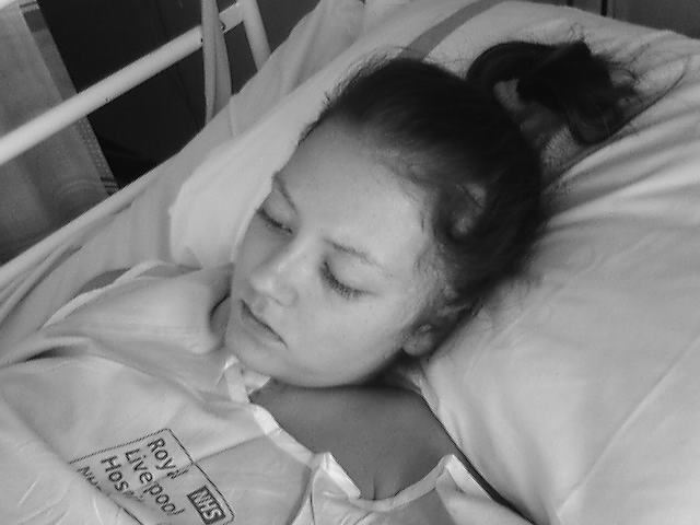 A black and white image of Christy, a white woman, she is laid in a hospital bed and looks sad and tired, she is wearing a hospital gown with the words NHS Royal Liverpool Hospital just visible