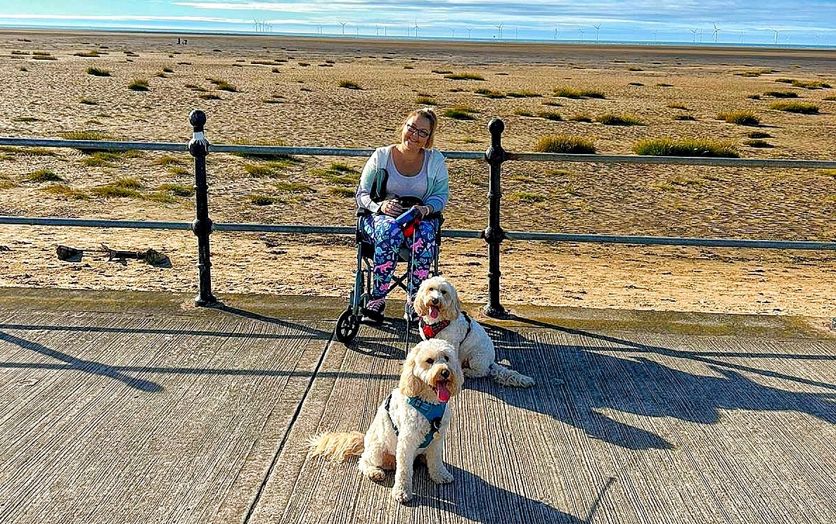 Christy is a white woman in her late 30s, she is sat in a wheelchair on a promenade by a beach with railing and sand and sea behind her, she is holding a dog lead attached to two light coloured dogs, possible labradoodles.