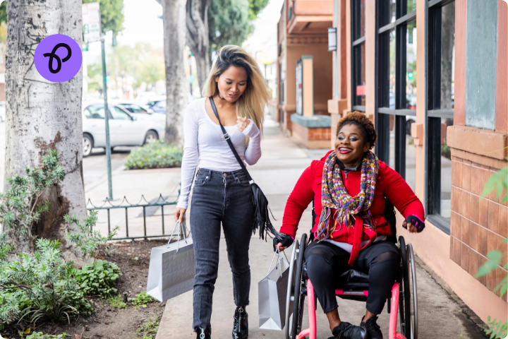 Two women shopping, they are going down a street, one is walking and one is a wheelchair user shopping, both are holding shopping bags for a purple Tuesday post about accessible shopping