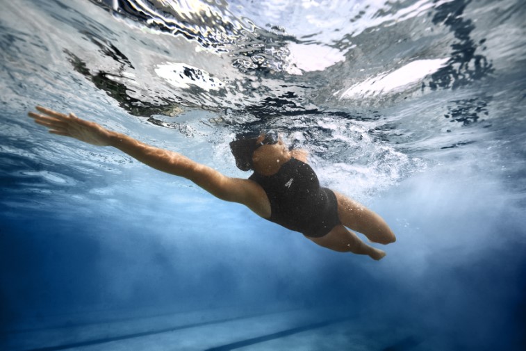 An underwater shot of a woman swimming, she is doing front crawl in a black swimsuit, swim cap and goggles with one arm outstretched and her face to the side for a speedo discount code with Purpl