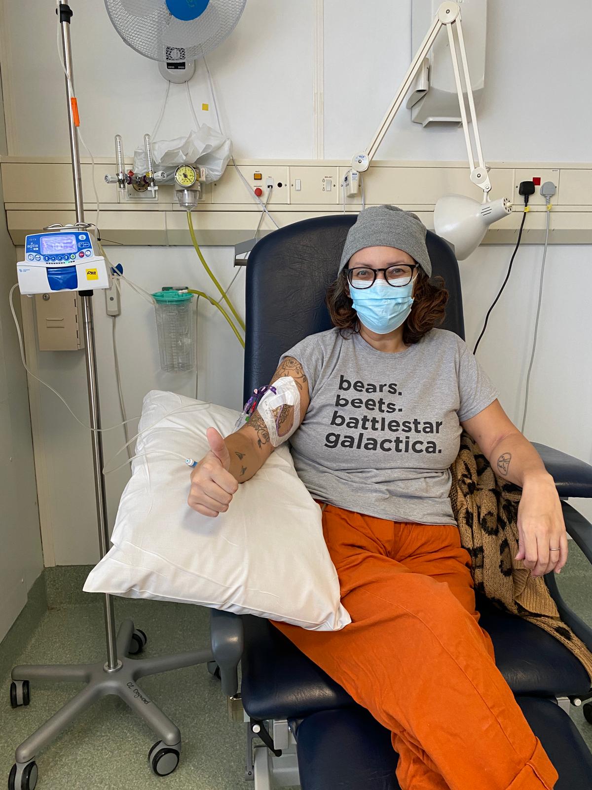 Sam Cleasby is a woman of Indian and British heritage, she is sayt in a hospital ward receiving an IV infusion, she is wearing a grey t-shirt that says bears beets Battlestar galactic and a face mask and is giving a thumbs up