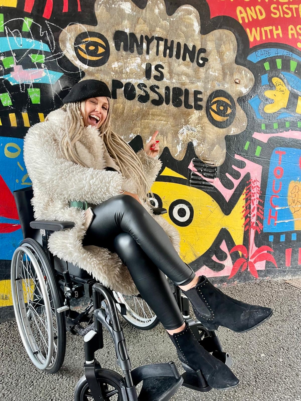 Anoushka is a white woman with long blonde hair, she is a wheelchair user and is wearing black leather look trousers, a white fluffy coat and a black beret, she is winking and pointing at a graffitied wall that says anything is possible