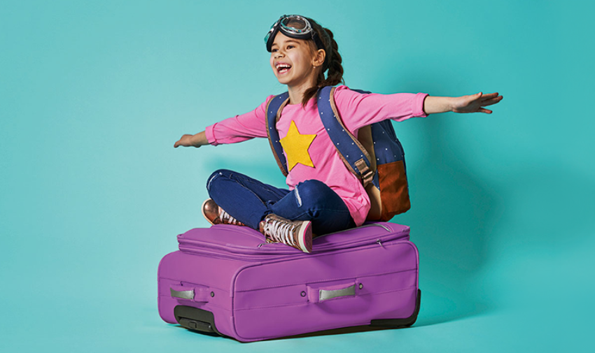 A young girl sits on a suitcase wearing a backpack and goggles pretending to fly with her arms outstretched