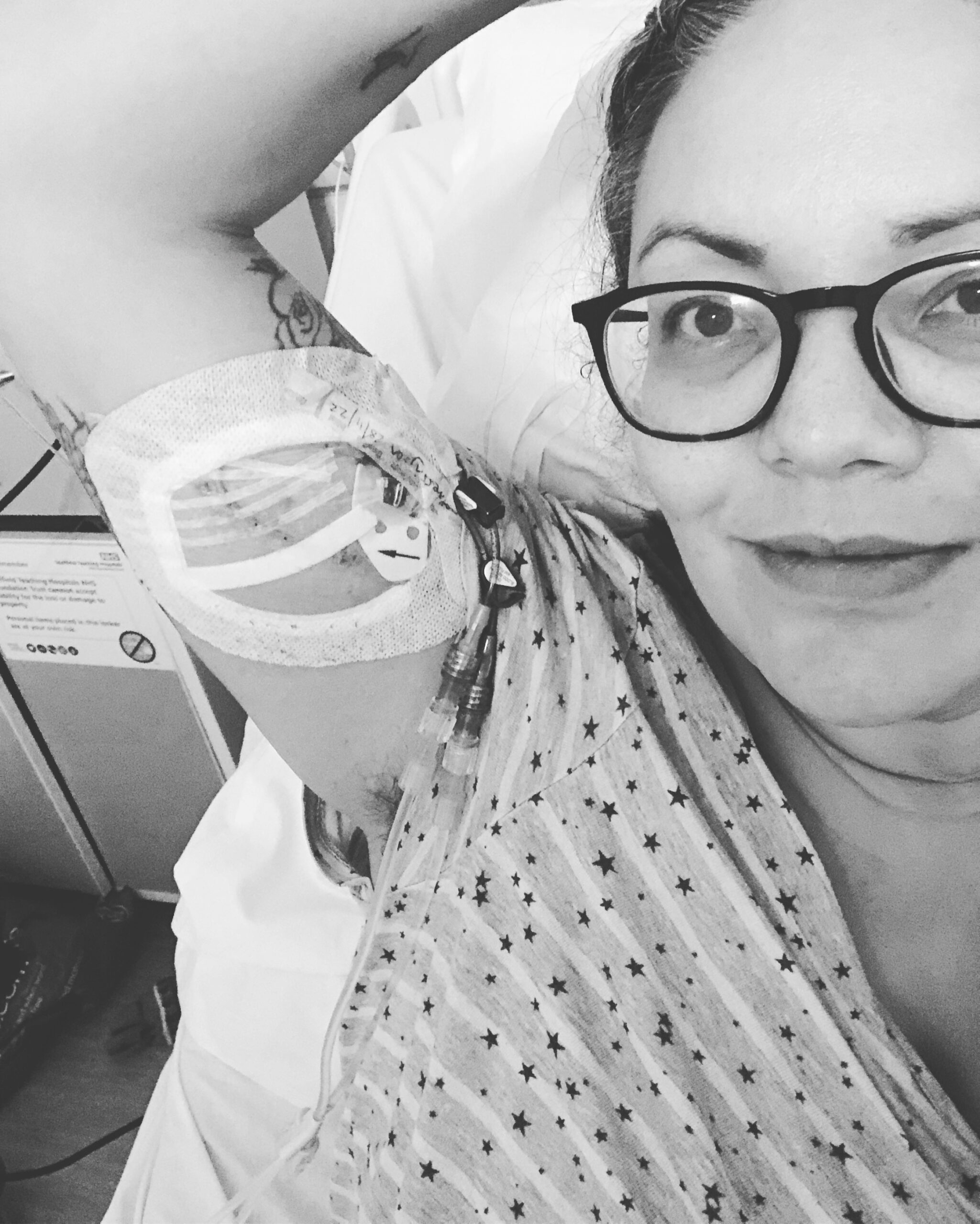 Black and white image of Sam Cleasby, a disability blogger and advocate who has IBD and a stoma, she is in a hospital bed and has a PICC line in her arm for TPN feeding and IV antibiotics 