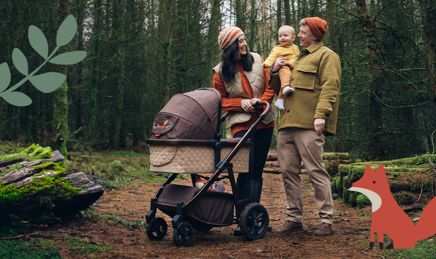 A male/female couple are walking in the woods dressed in outdoors gear and have a pram and a baby in the mans arms, there are graphically drawn images on a fox and a leaf