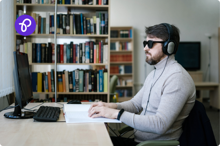 A visually impaired man is wearing dark glasses and headphones and is sat studying at a desk and computer in a uni library wearing a grey turtleneck jumper