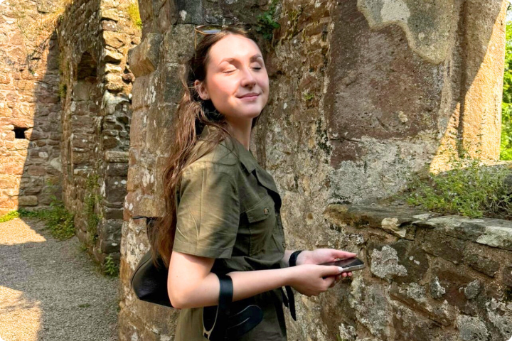 A young woman with long brunette hair and wearing an olive-green outfit is next to a stone wall of ancient ruins, smiling softly with her eyes closed in the sunlight. She holds a phone and has a black crutch on her forearm. Surrounding area is filled with rustic stone and some greenery.