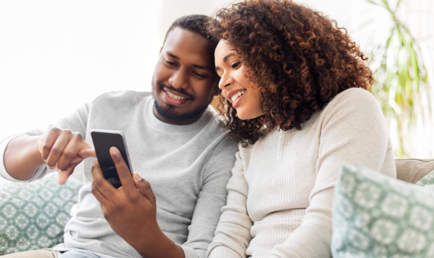 A couple sit on a sofa smiling, they are looking at a phone and the man is pointing