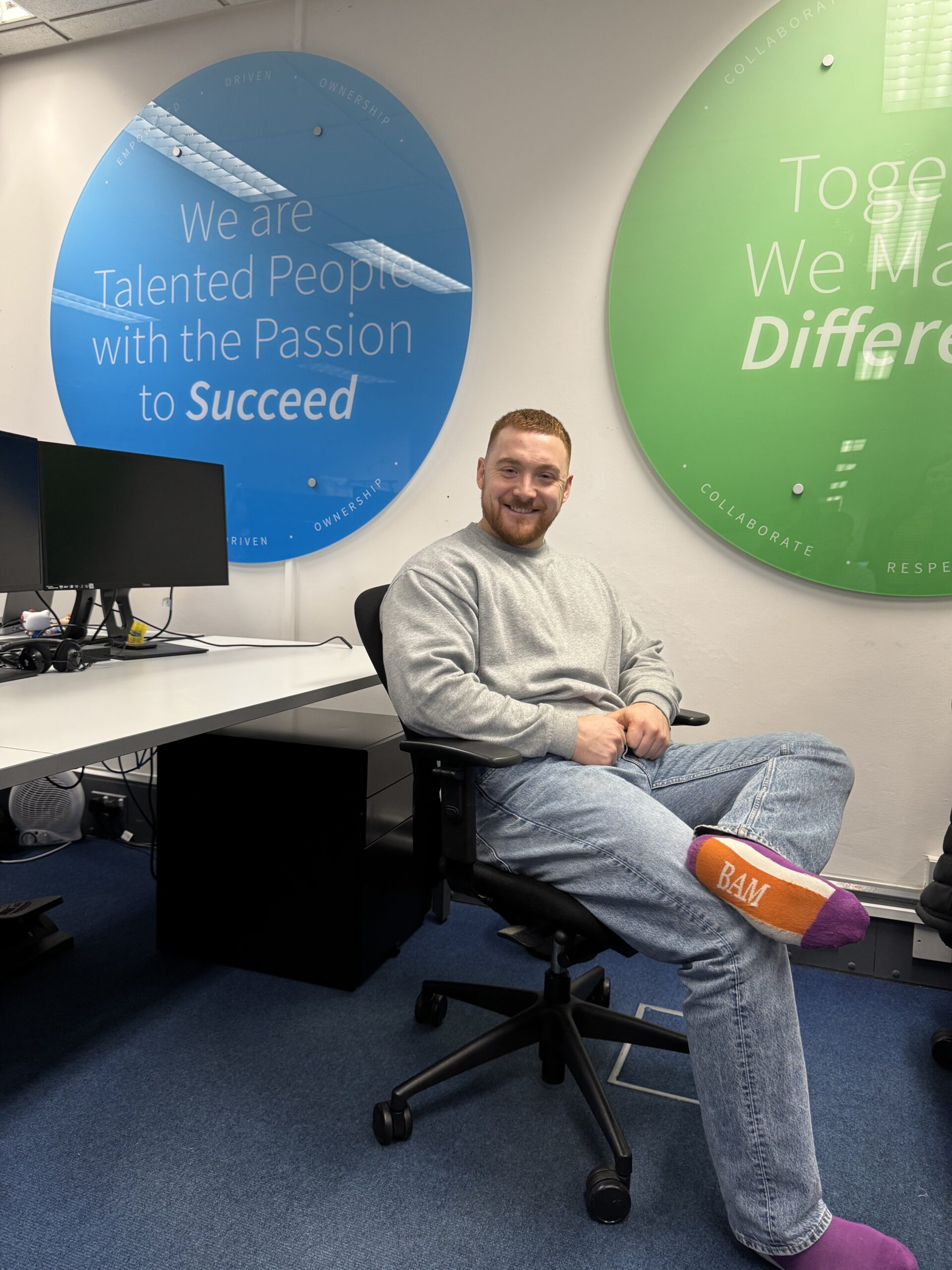 A man sits in an office wearing jeans and purple and orange bam socks celebrating Purple Sock Day for the International Day of Disabled People 