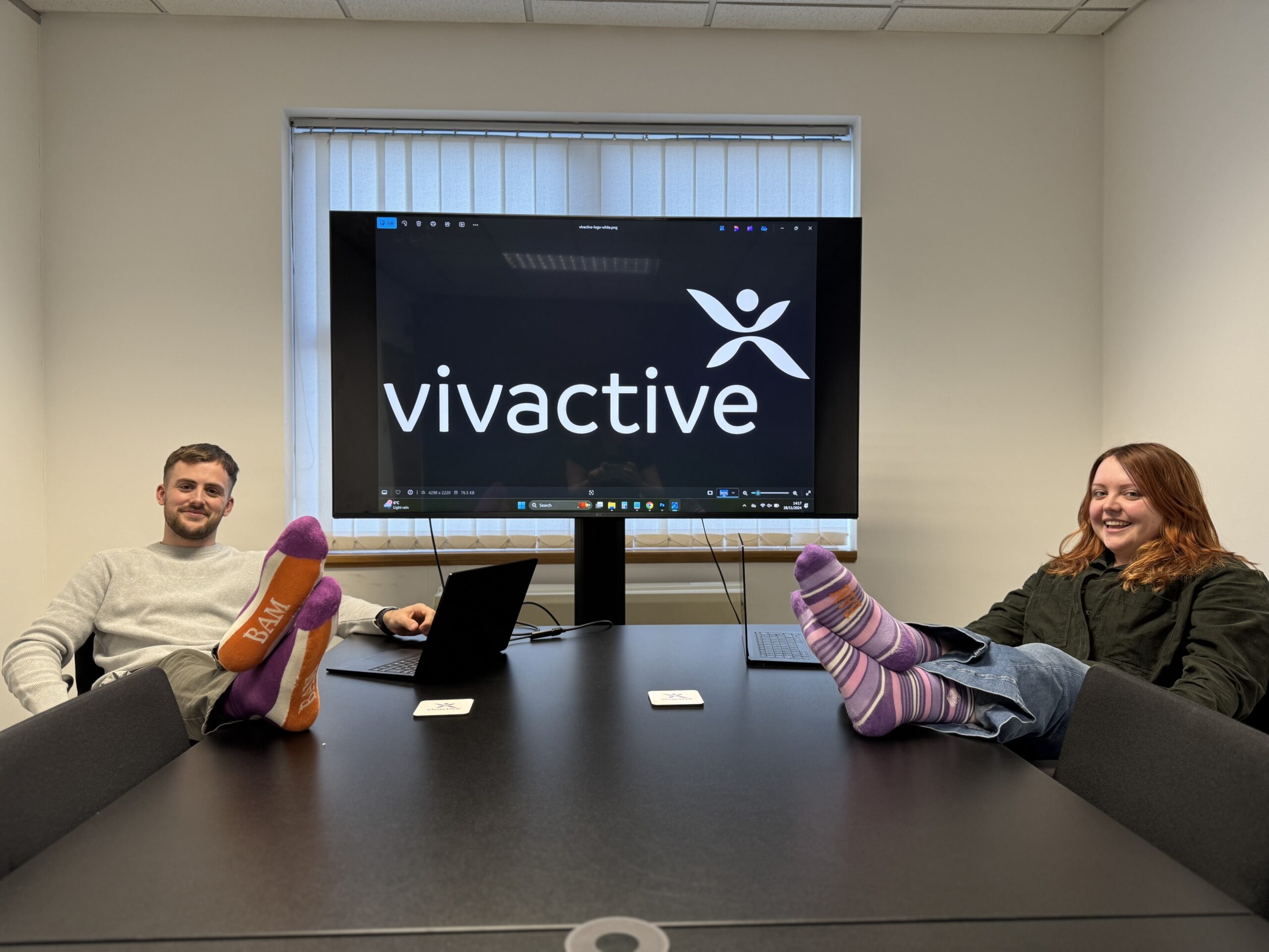 A man and a woman pose with their feet on a table in an office with a big screen with the Viactive logo on it, they have Bam socks and are celebrating Purple Sock Day for the International Day of Disabled People 