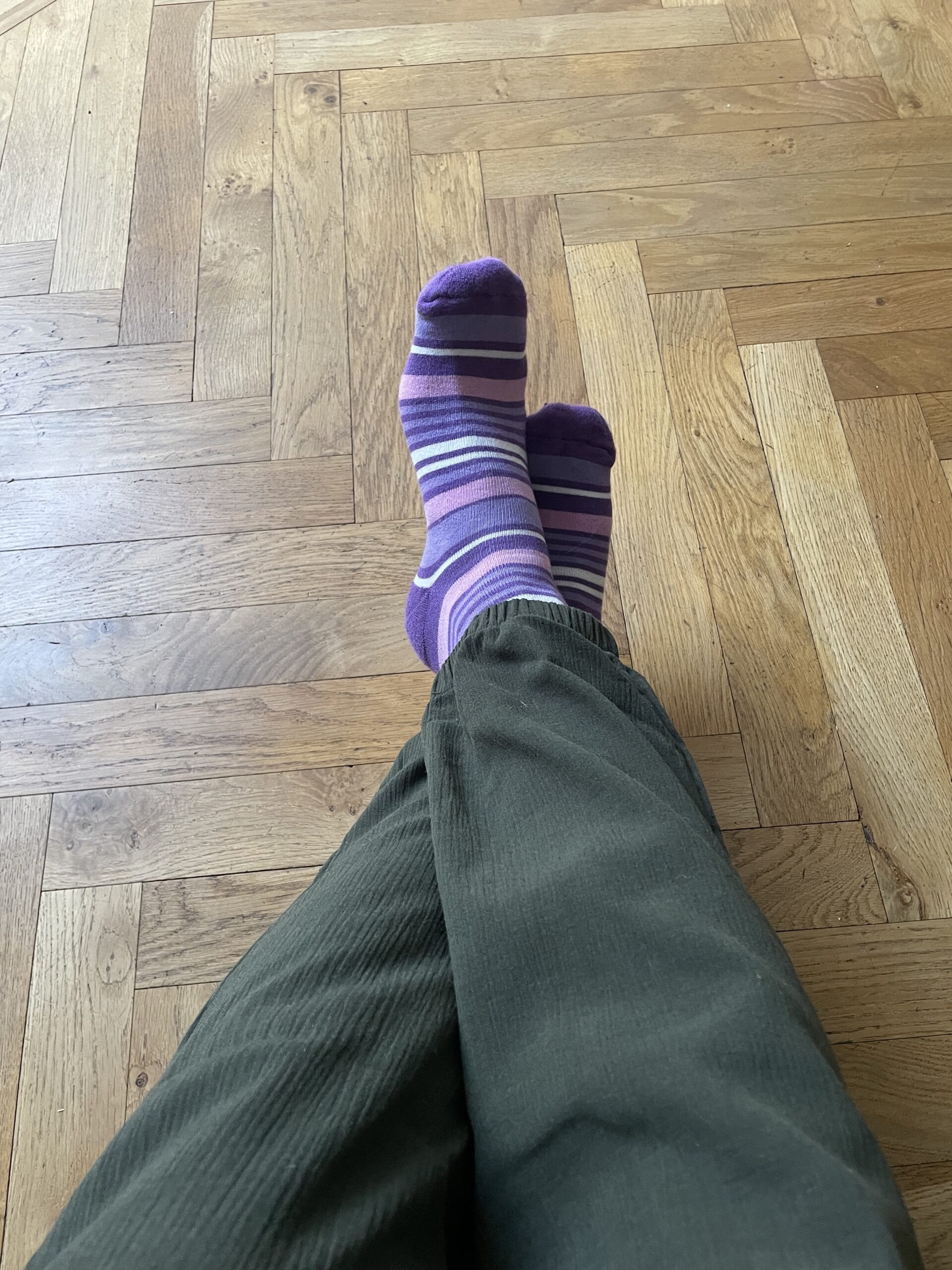 A pair of legs, crossed at the ankle wearing olive coloured trousers and purple stripy socks for purple sock day 2024