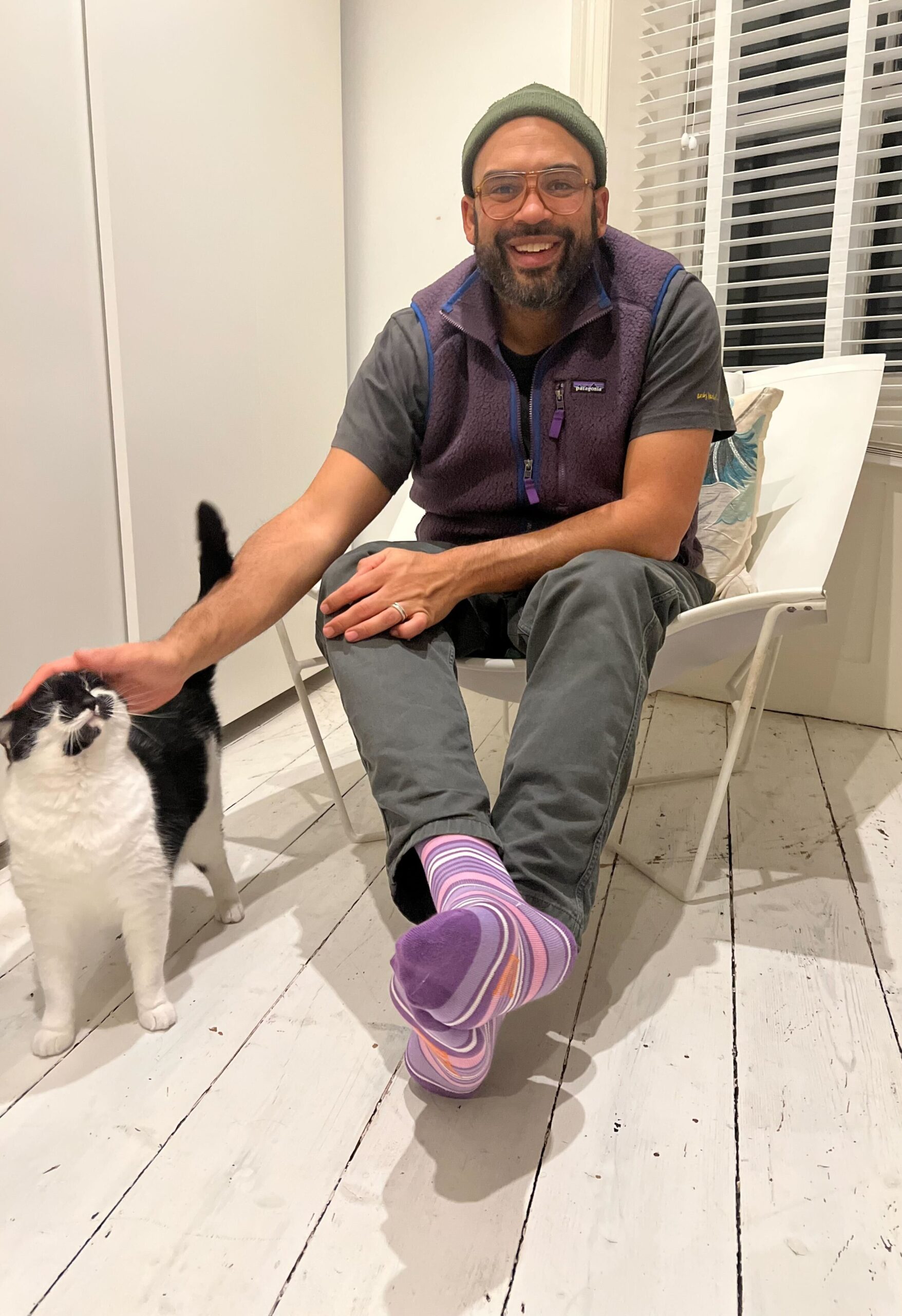 A black man wears glasses and a beanie hat, casual clothes and purple strip socks, he is sat in a chair and stroking a black and white cat and smiling, he is celebrating Purple Sock Day for the International Day of Disabled People 