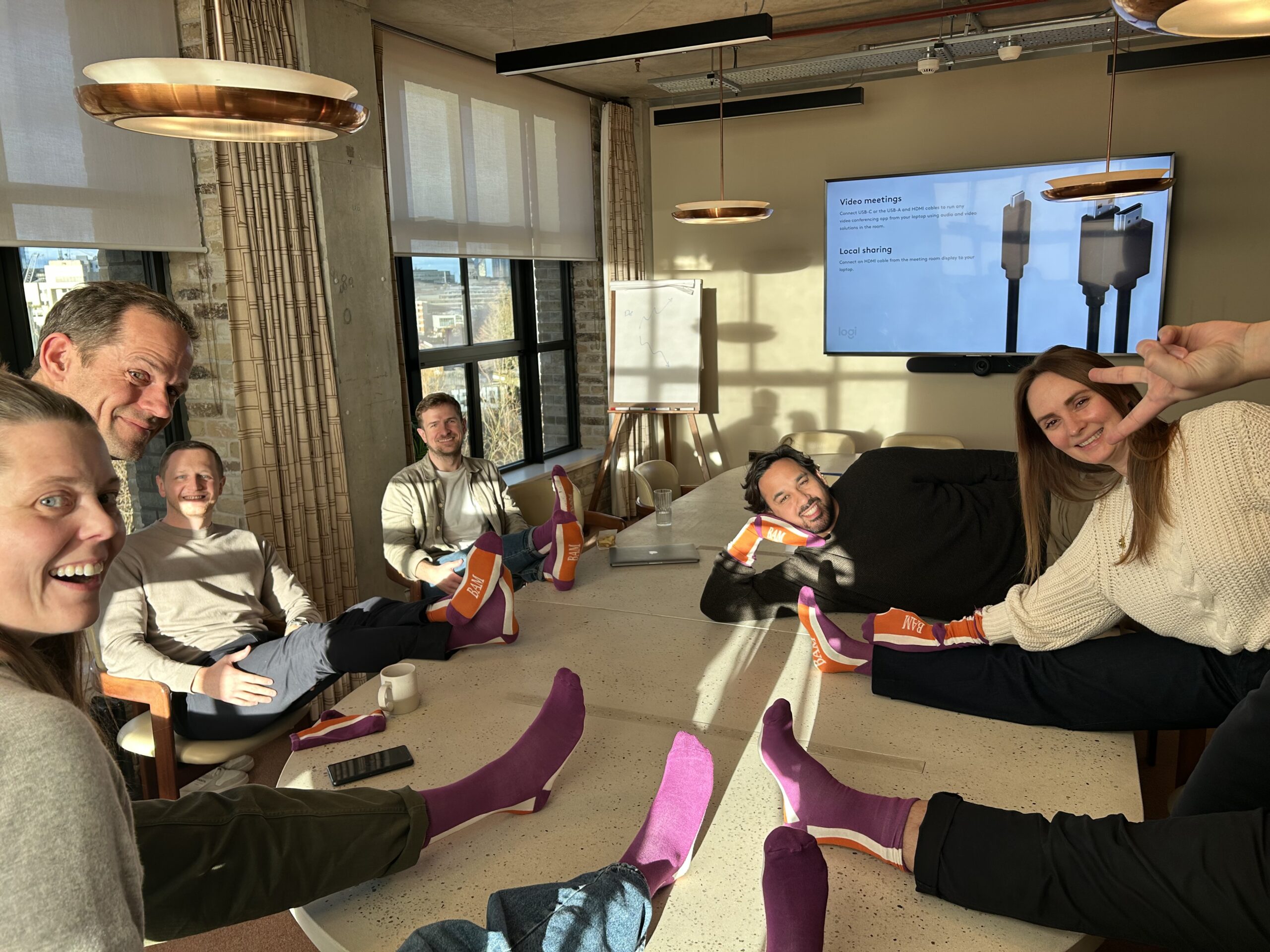 A team of staff hang our around a meeting table in an office celebrating Purple Sock Day for the International Day of Disabled People 