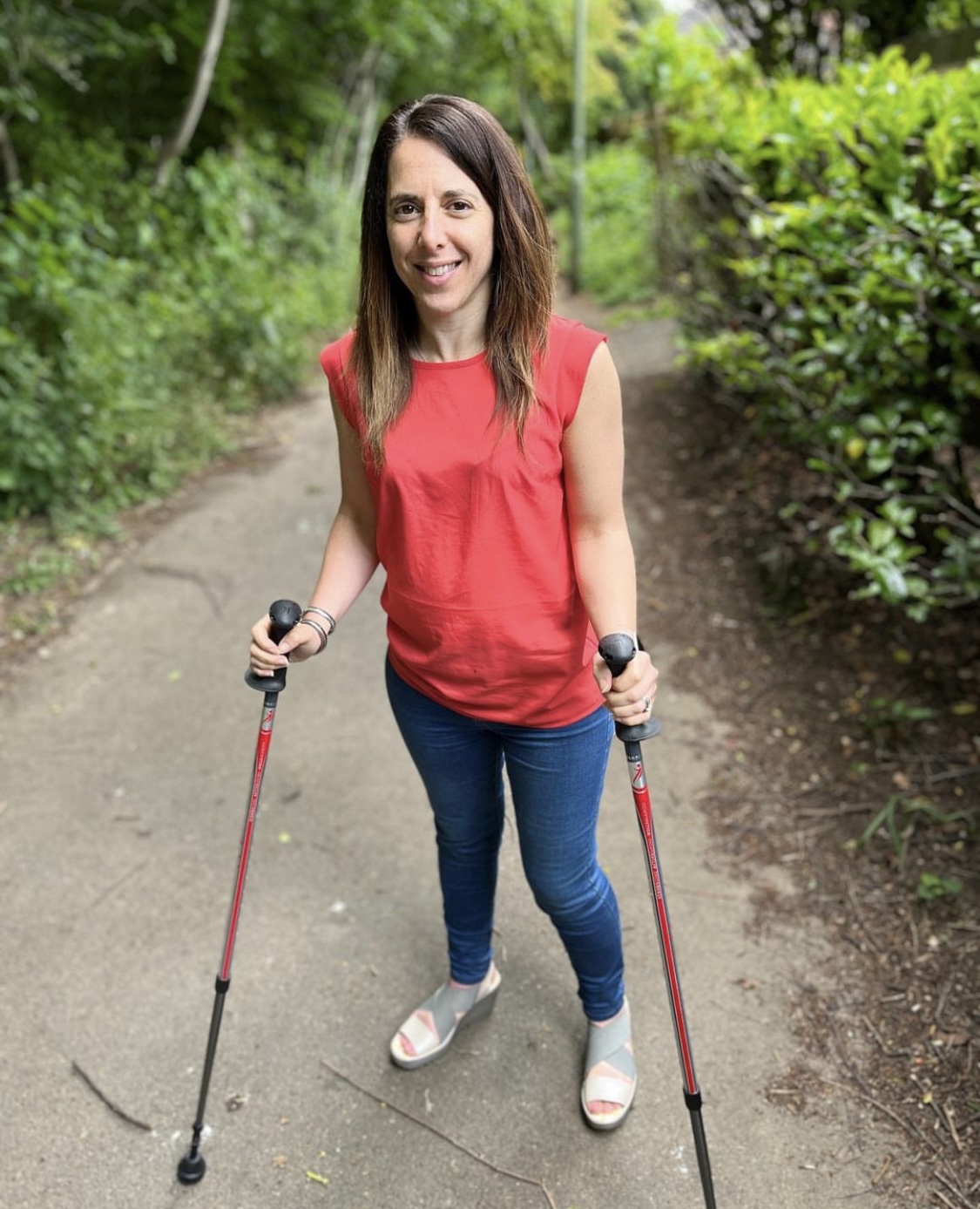 Georgina is a white woman with long dark straight hair, she is wearing a sleeveless red top and jeans and is walking outdoors on a path with green trees and bushes round her and is using two walking sticks