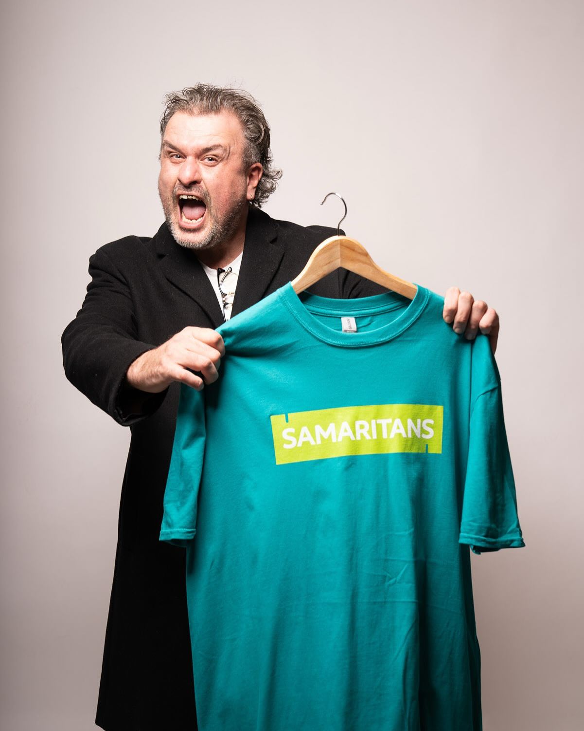 Jon McClure is a white man in his 40s, he has greying hair and a gold front tooth, he is holding up a green samaritans t-shirt and has his mouth open as though he is shouting 