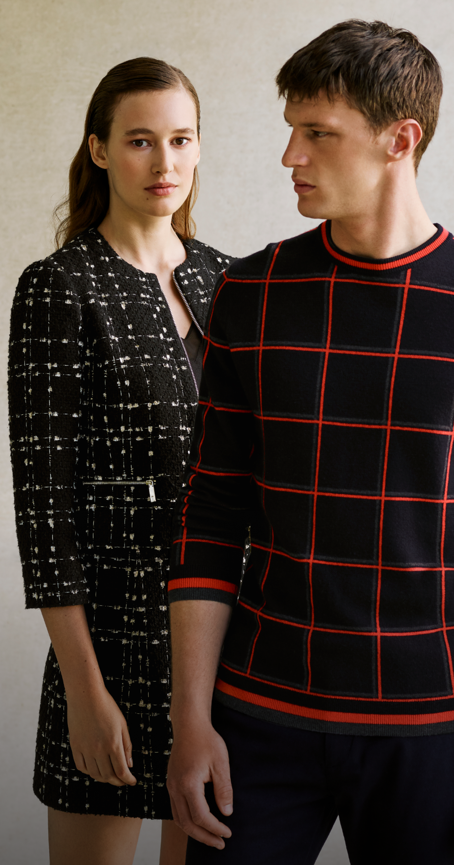 a man and woman pose on white background, she looks to camera and is wearing a black and white suit and he looks to the right wearing a black jumper with red squares on it