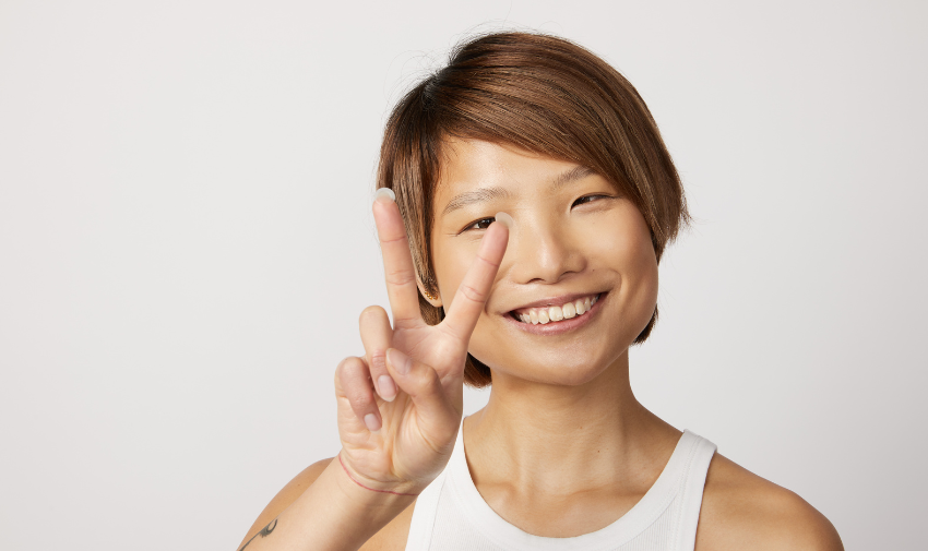 An young asian woman with short brown hair and natural beauty and skin smiles to camera and holds her fingers up in a peace sign