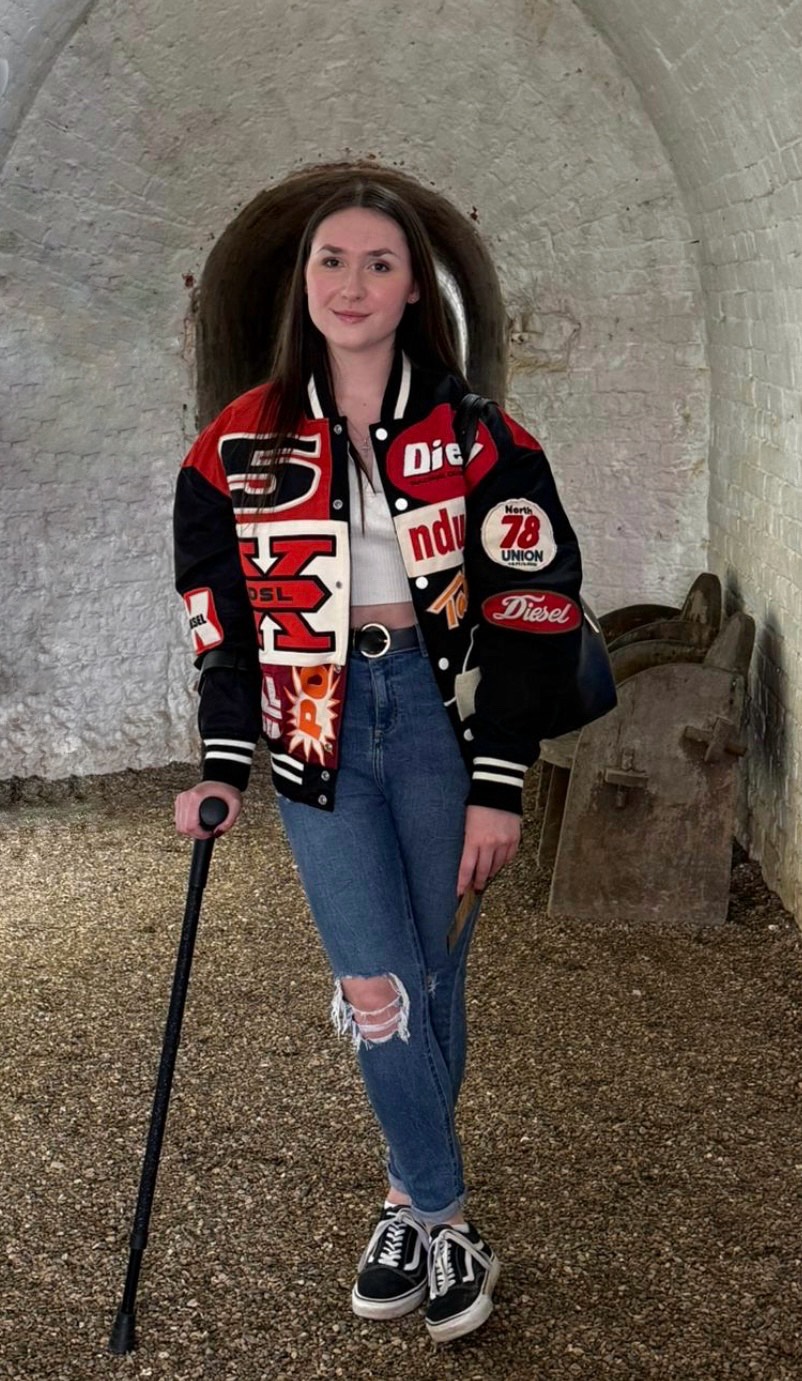 A young woman with long brunette hair stands in a white stone archway, wearing a varsity jacket, ripped jeans, and black-and-white trainers. She is using a black crutch on one side, with an old wooden bench in the background.