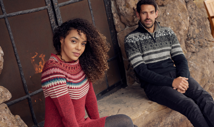 a man and woman pose outdoors sat on a stone wall with a lead window behind them, she is mixed race and has curly dark hair and a red winter jumper, he wears a dark christmas jumper and has short dark hair and a cropped beard and is looking at the woman