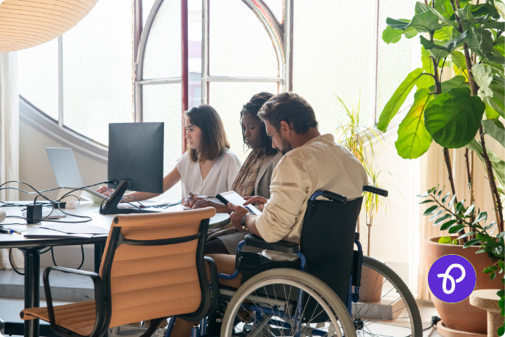 A group of three people are sat at a desk in an office, there are two women and a man, the first woman has shoulder length brown hair, the second lady is black and wears a beige jacket and the man s white, has a beard and is a wheelchair user