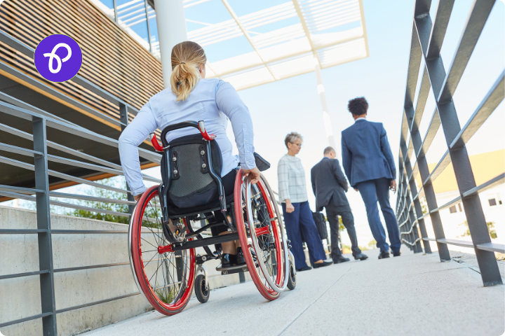 An outside scene outside an office block, there is a white woman with blonde hair who is a wheelchair user, she is wearing a blue blouse and her wheelchair has red wheels and she is pushing herself up a ramp