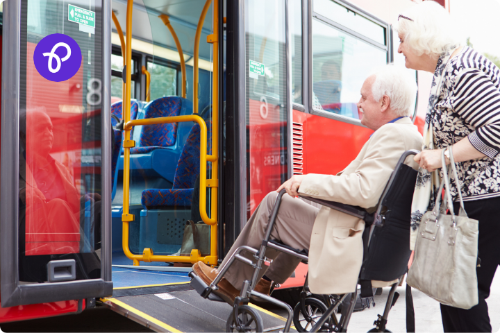 A couple getting on a red bus using a ramp, a man has white hair and wears a suit and is a wheelchair user and a woman with grey hair and a stripy top is pushing the wheelchair