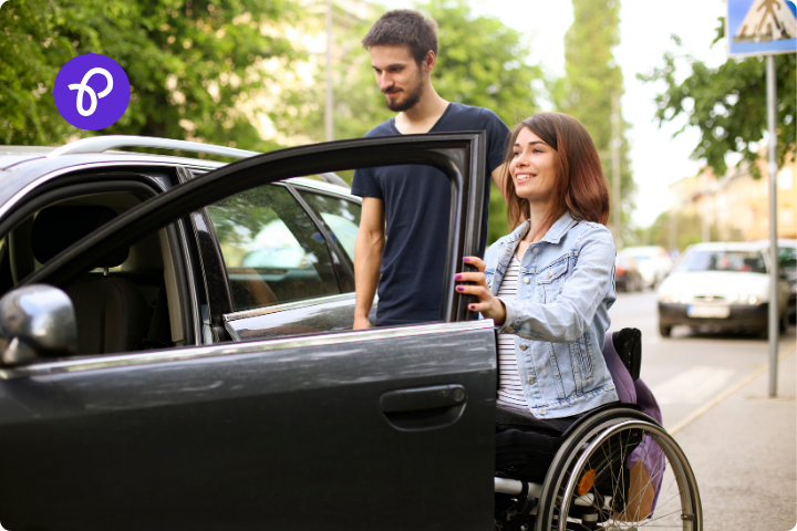 A woman is a wheelchair user and is getting into a dark car, she is wearing a denim jacket and is with a young white man with a beard and they are on a street