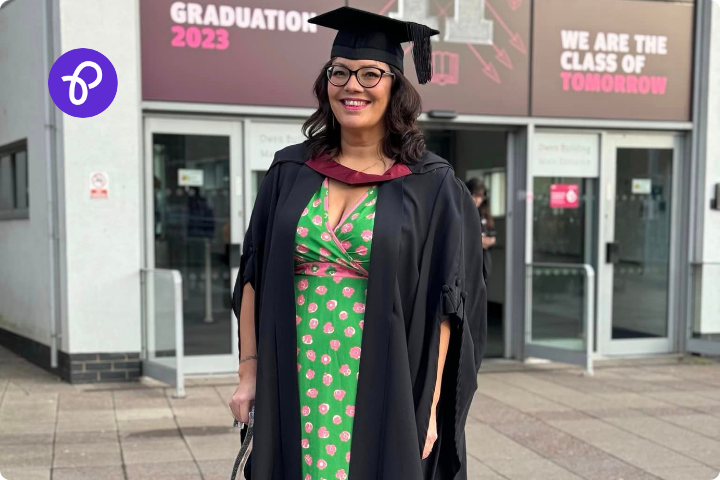 Sam Cleasby is a brown woman in her early 40s, she is in a graduation gown and cap and stands in front of a university building with text saying Graduation 2023 and We are the class of tomorrow she is wearing a green and pink dress and glasses and she uses a mobility cane