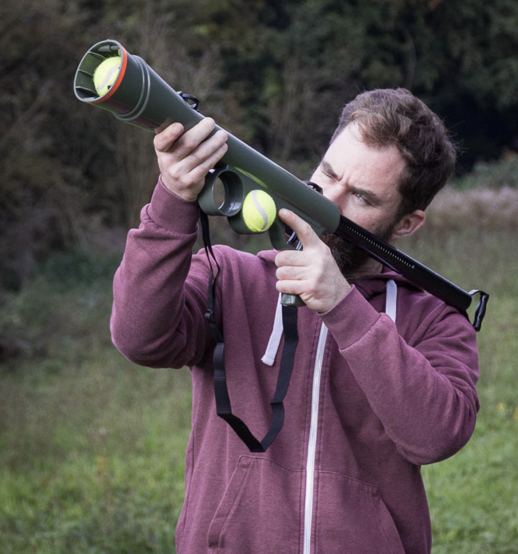 A man is holding a ball launcher for dogs over his shoulder outdoors, he is white and around 40 with a maroon hoodie on.