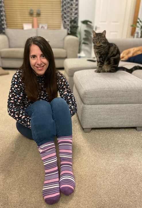 Georgina is a white woman in jeans and stripy socks, she has long dark hair and is sat in a living room with a cat celebrating Purple Sock Day for the International Day of Disabled People 